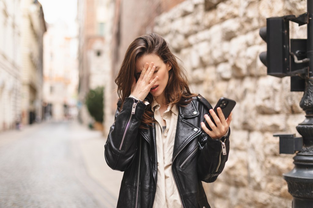 Annoyed woman on the street covering her face with one hand while holding a cell phone in the other