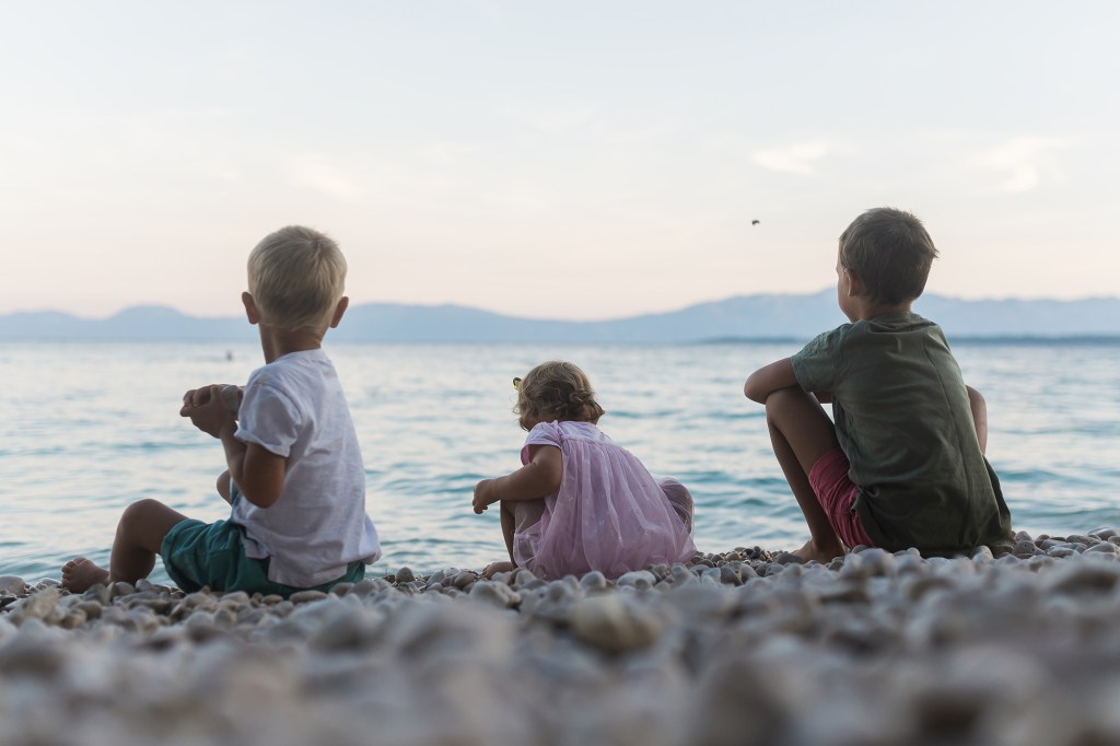 Three children, two boys and a girl, sitting on a pebble beach in the evening, throwing stones into the sea