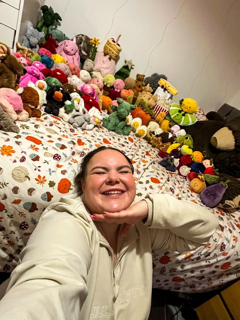 The woman named Megan lying on a bed surrounded by her extensive collection of Jellycat plush toys