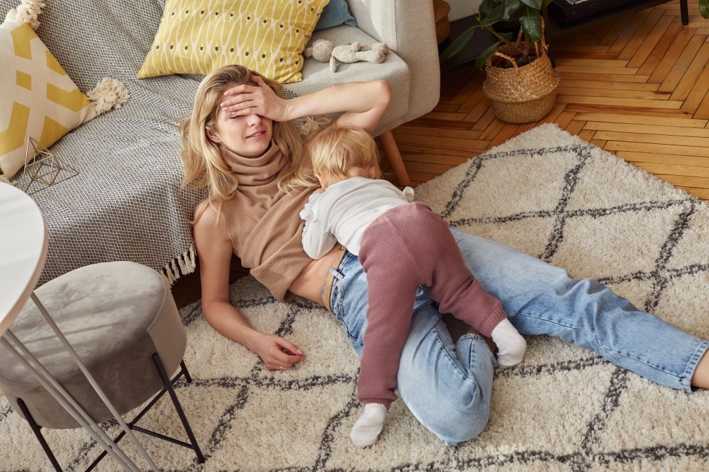 Caucasian blonde woman with jeans sitting in the carpet, breastfeeding her baby in a bright, Scandinavian style room