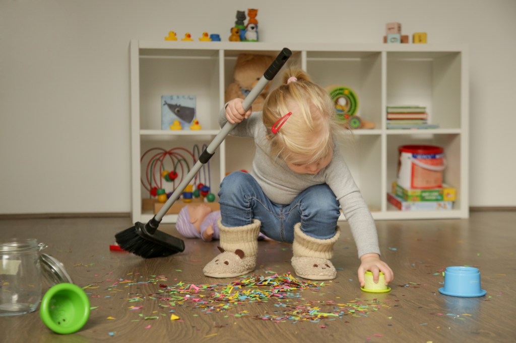 Little adorable blonde girl who cleans the colorful confetti from the floor with a broom